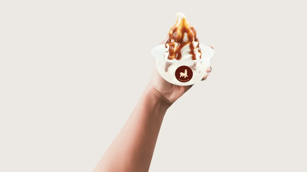 Soya soft serve: hand holding up an Alpacco cup of soya soft serve with brown syrup drizzle