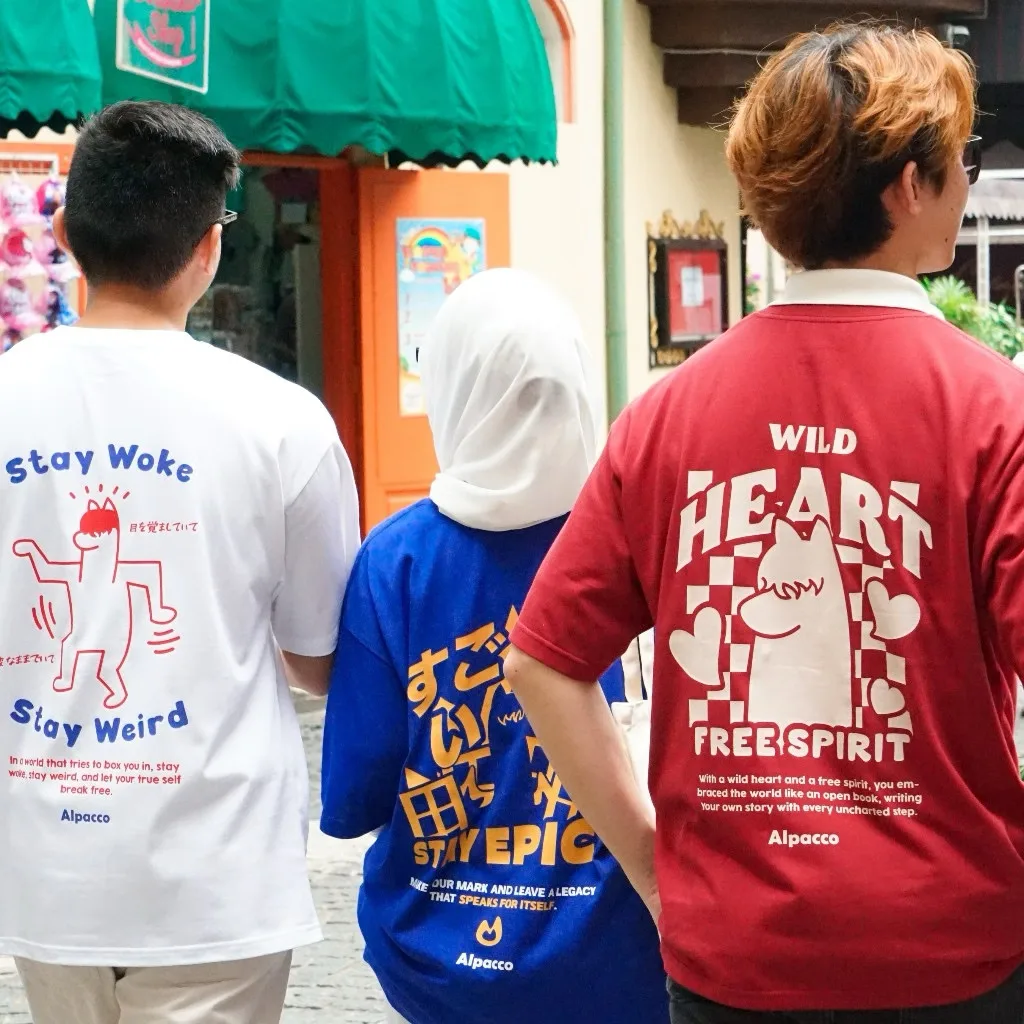 Three friends seen from behind on a Malaysian street wearing Alpacco graphic tees with Stay Woke Stay Weird, Stay Epic and Wild Heart Free Spirit back prints